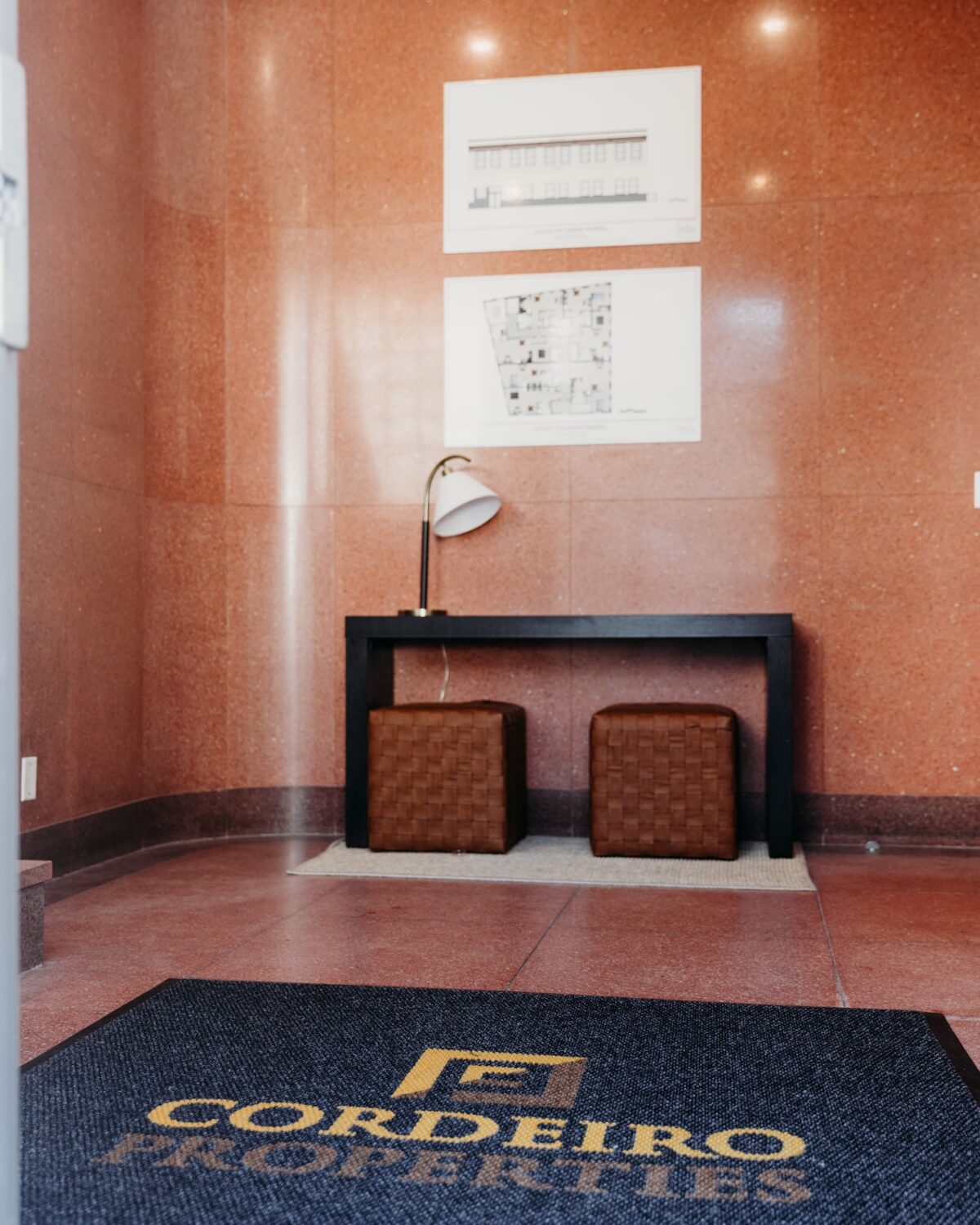 Interior of a lobby featuring a black table, two brown stools, a lamp, and a welcome mat with "Cordeiro Properties.