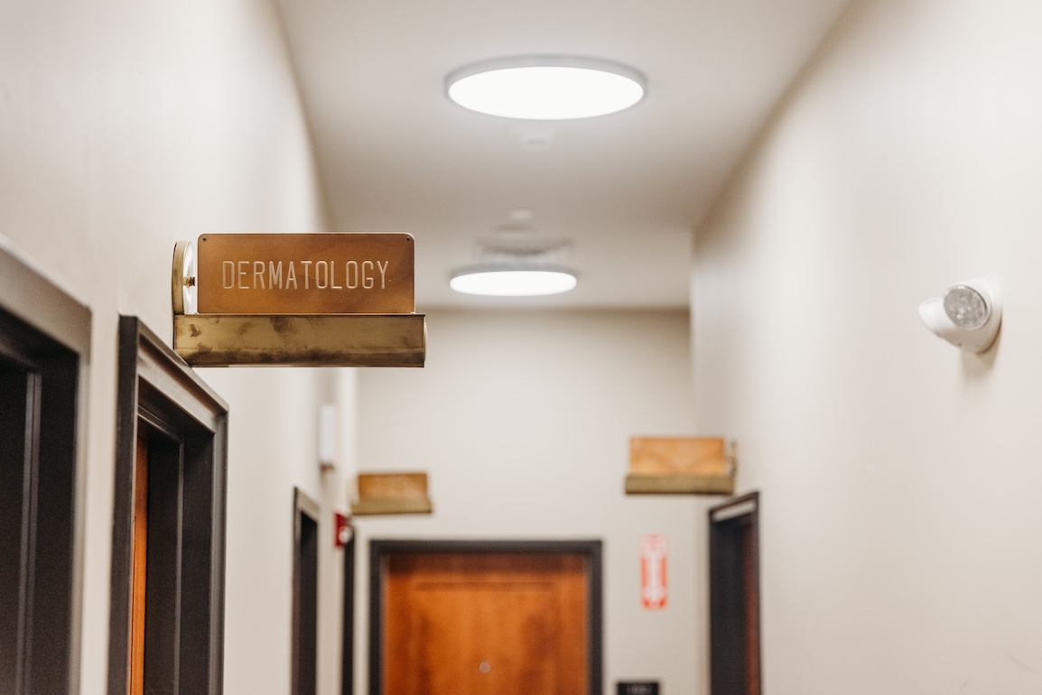 Sign for "Dermatology" hangs above a hallway with closed doors and overhead lighting.