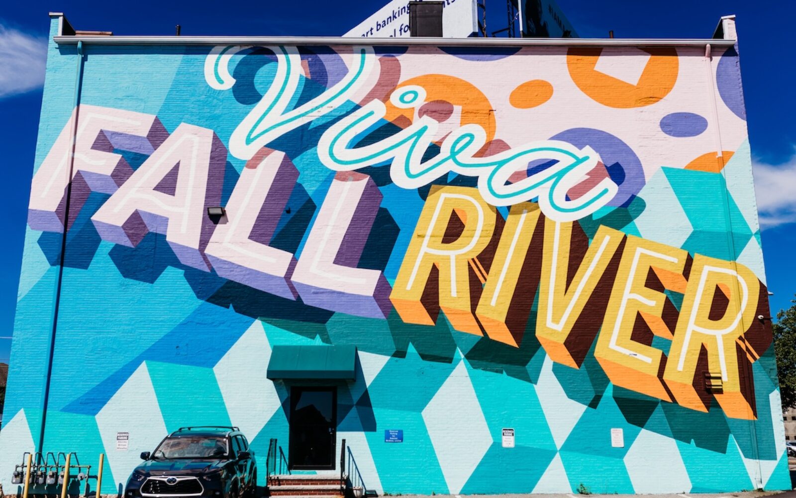 Colorful mural reading "Viva Fall River" on a building, with geometric patterns and a parked SUV below.
