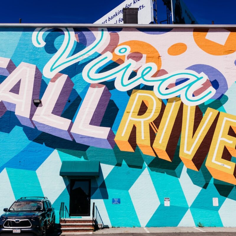 Colorful mural reading "Viva Fall River" on a building, with geometric patterns and a parked SUV below.