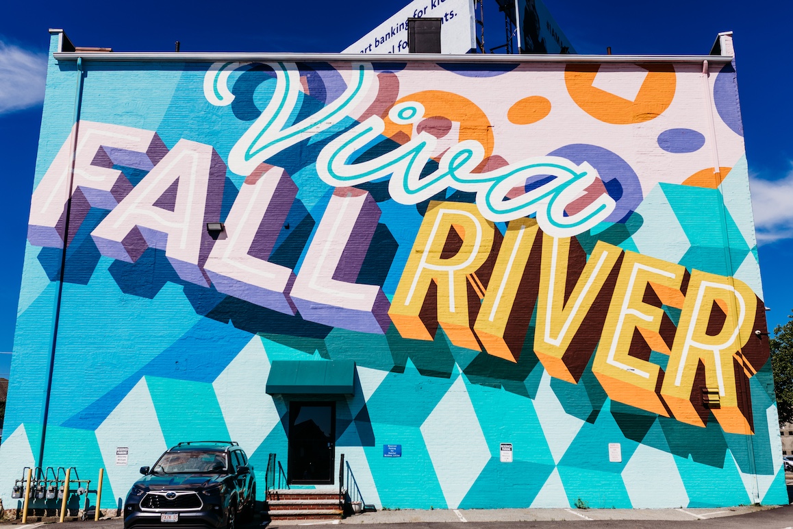 Colorful mural reading "Viva Fall River" on a building, with geometric patterns and a parked SUV below.