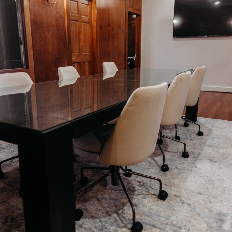 A modern conference room with a glass table and several beige rolling chairs. A TV is mounted on the wall.
