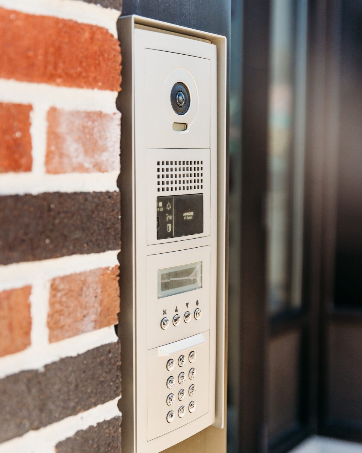 A wall-mounted intercom system with a camera and keypad, next to a brick wall.