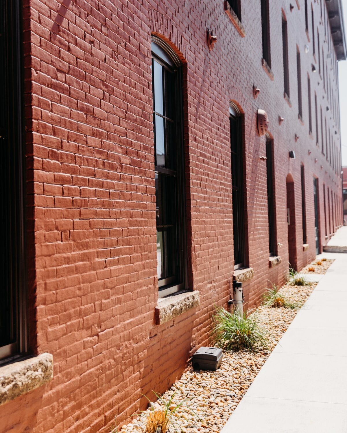 Red brick building exterior with windows, bordered by a gravel path and small plants.