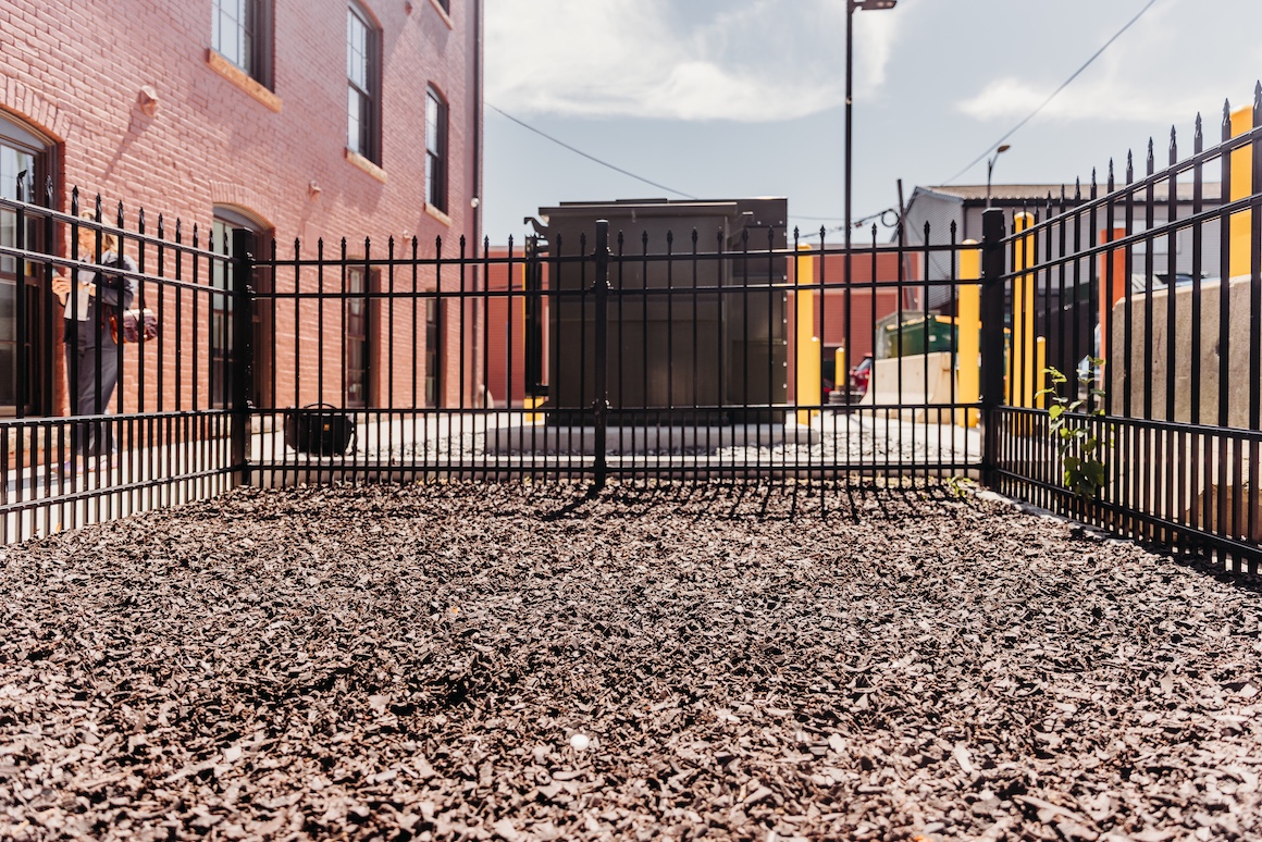 A fenced area with dark mulch, a utility box in the background, and a person standing near a building.