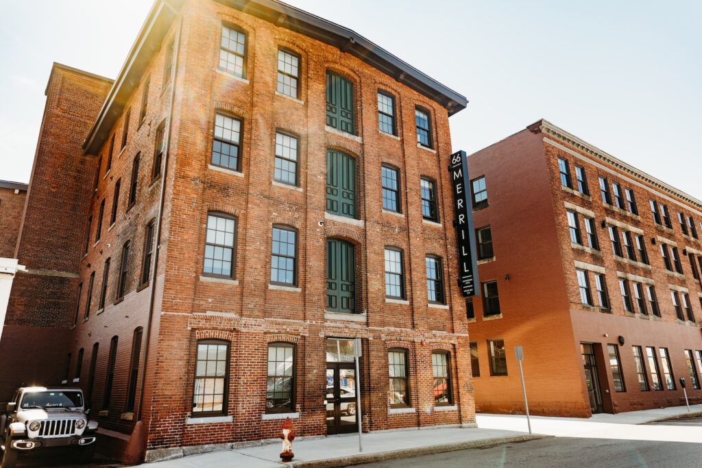 Brick building with large windows and a sign reading "66 MERRILL." A parked vehicle is visible nearby.