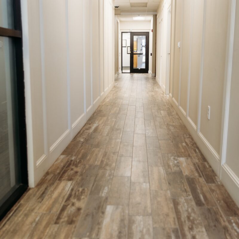 A long, narrow hallway with wooden flooring and light-colored walls, leading to a glass door at the end.