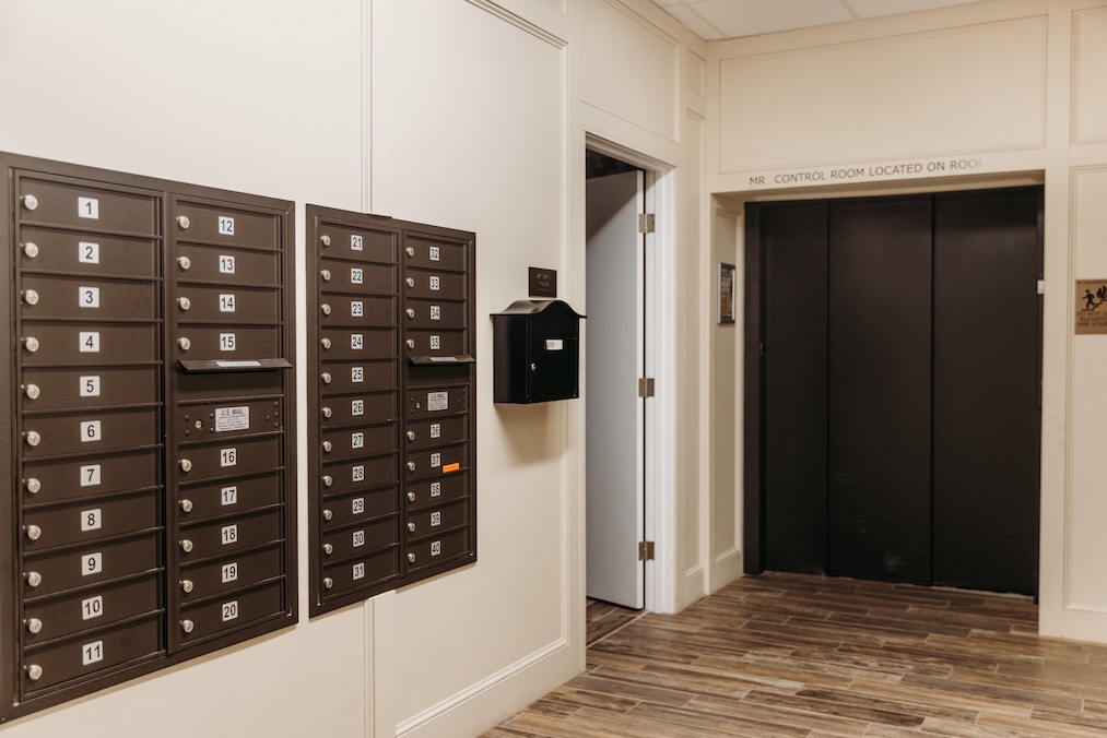 Mailboxes line the wall beside an elevator in a hallway. A door leads to another room.