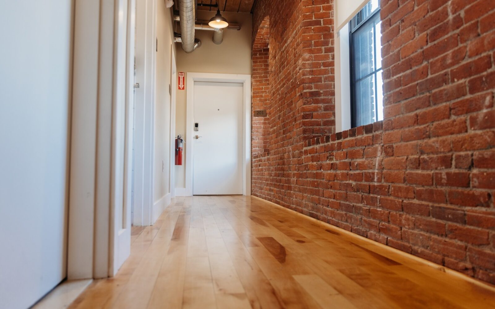 A hallway with wooden flooring, exposed brick walls, and a door at the end.