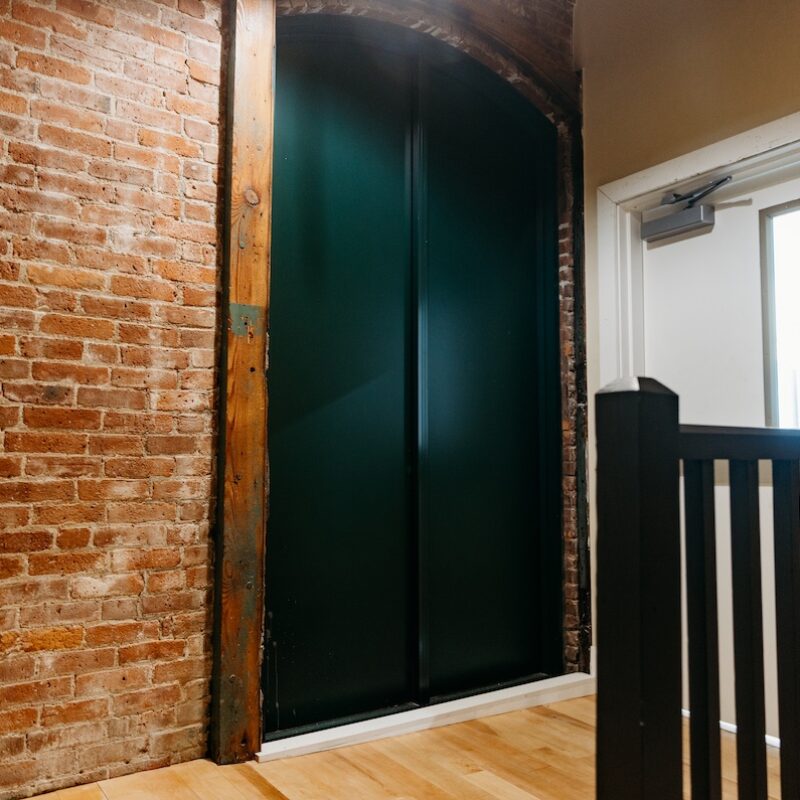 A hallway with exposed brick walls, a green double door, and a white door leading outside.