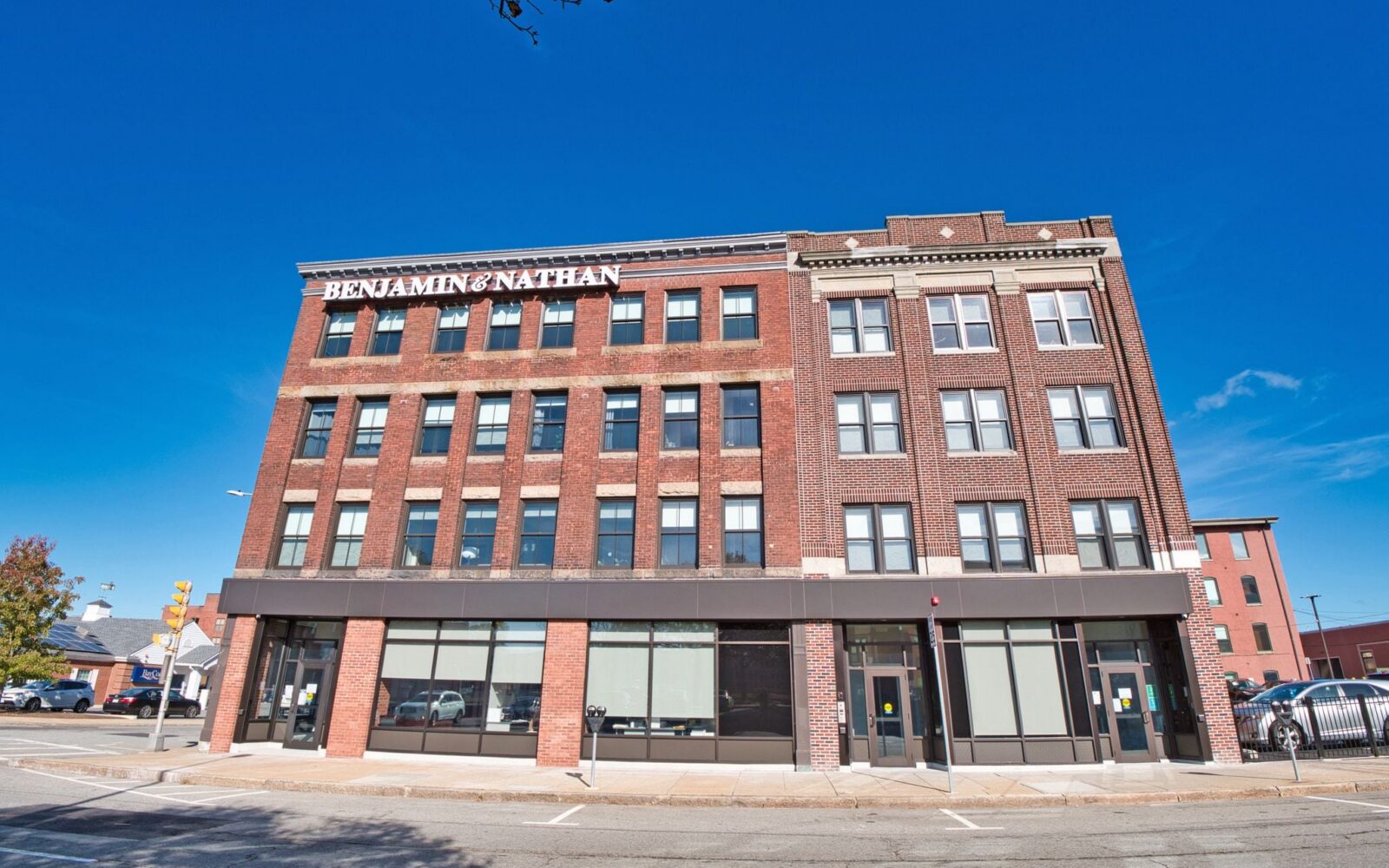 Brick building with "BENJAMIN & NATHAN" sign, large windows, and clear blue sky. Street view with parked cars.