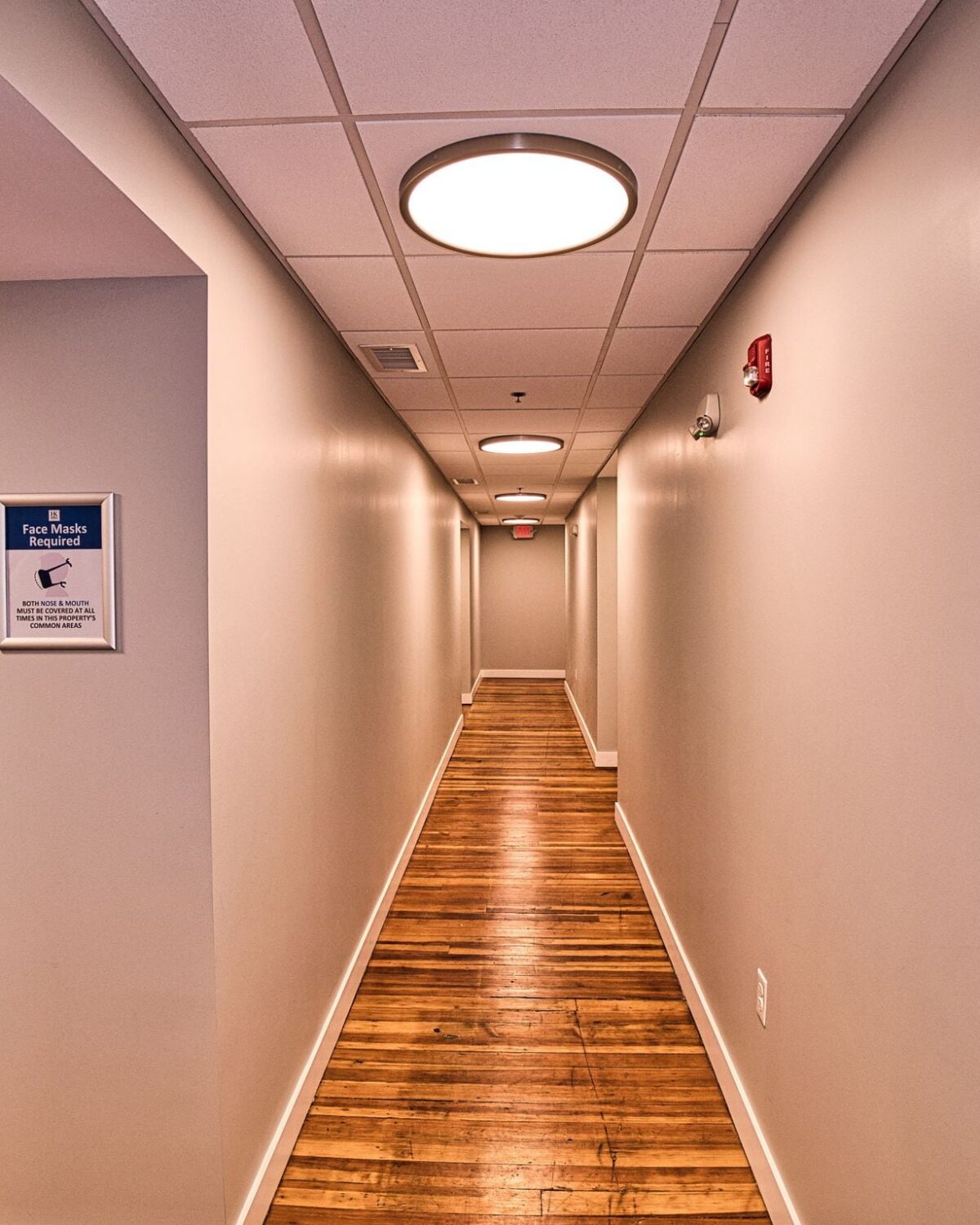 Long hallway with wooden floors, elevator doors, and a sign indicating face mask requirements. Exit sign visible.