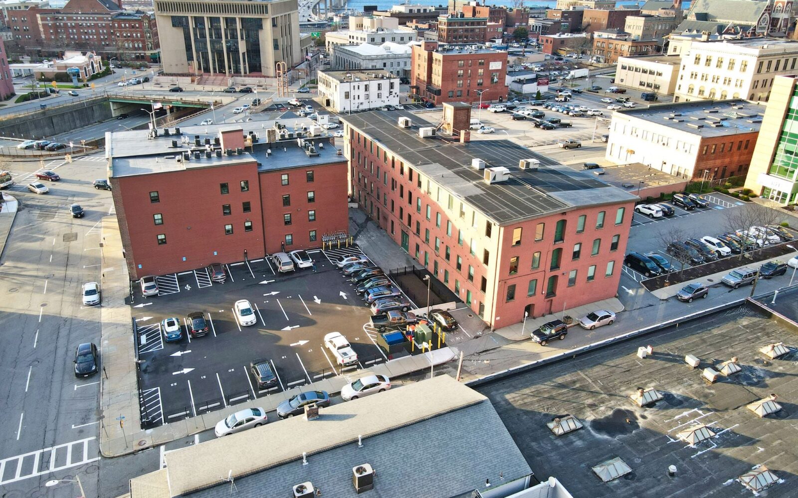 Aerial view of a parking lot surrounded by buildings, with cars parked and a bridge in the background.