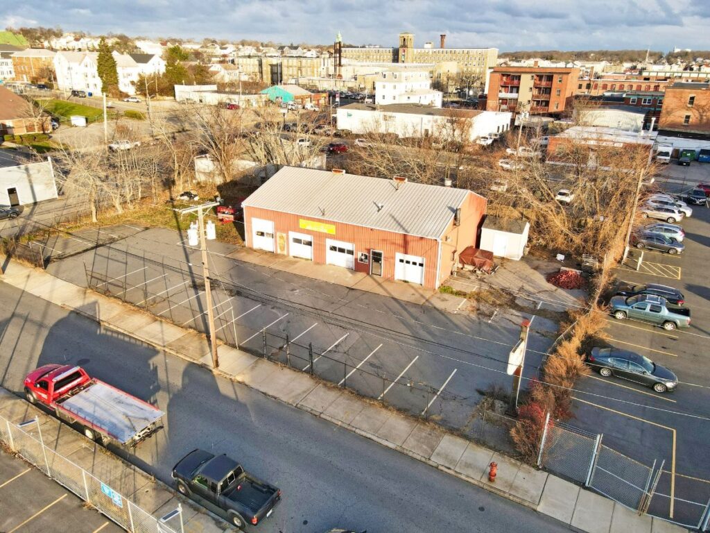Aerial view of a red building with a metal roof, surrounded by parking lots and trees. Two trucks on the road.