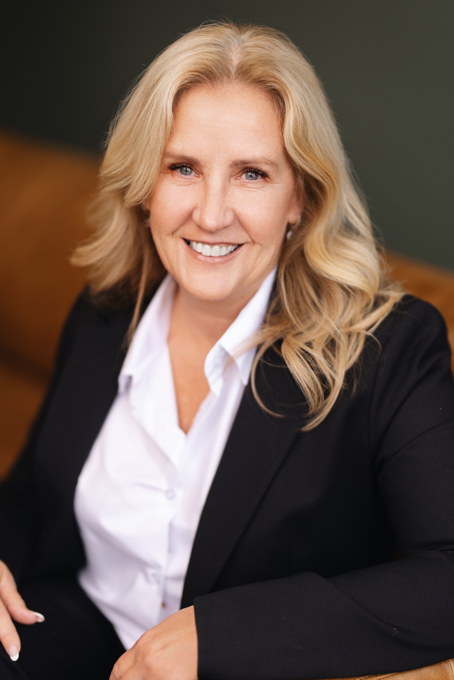 A smiling woman with long blonde hair wears a black blazer and white shirt, seated on a couch.