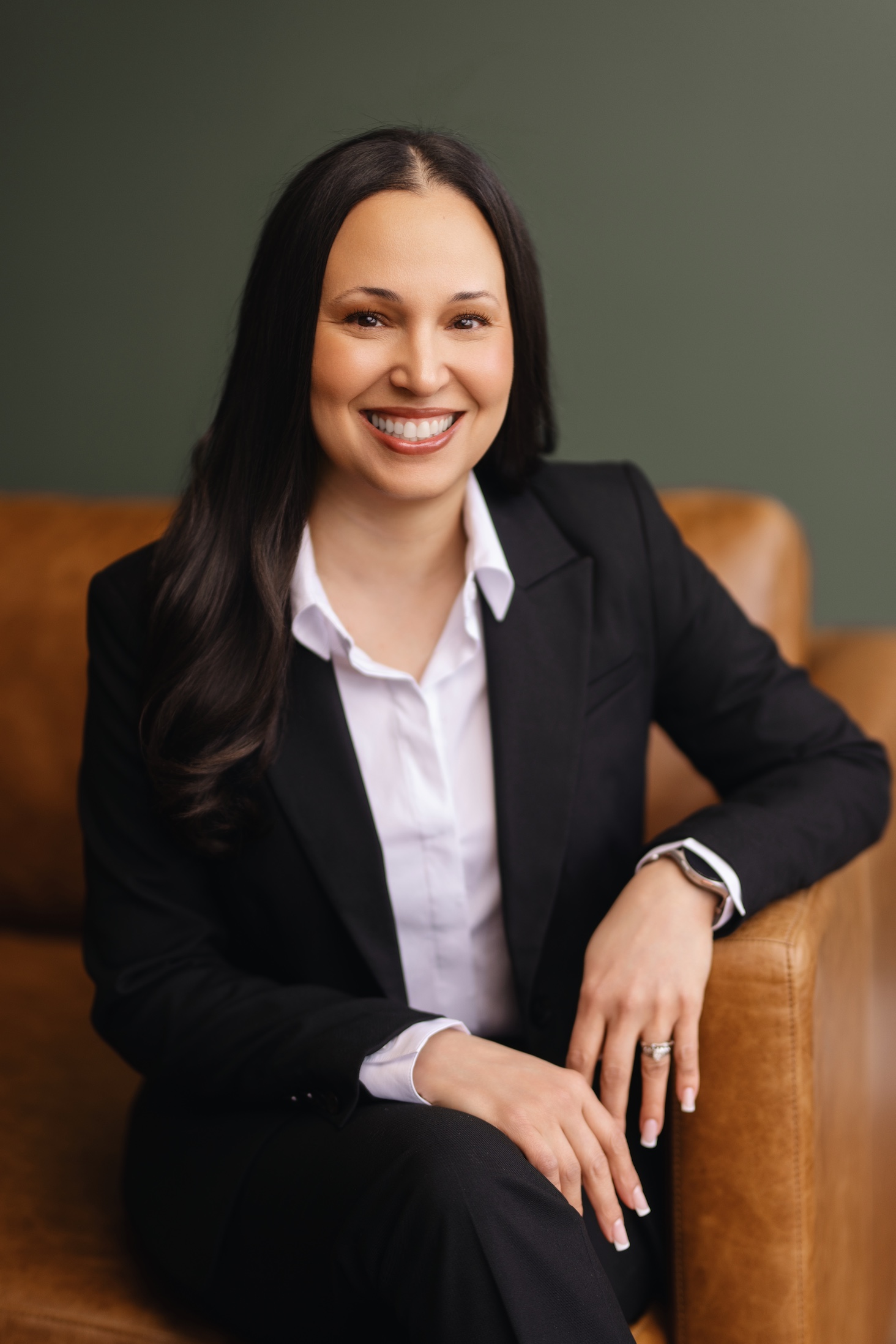 A woman in a black suit smiles while sitting on a brown leather couch.