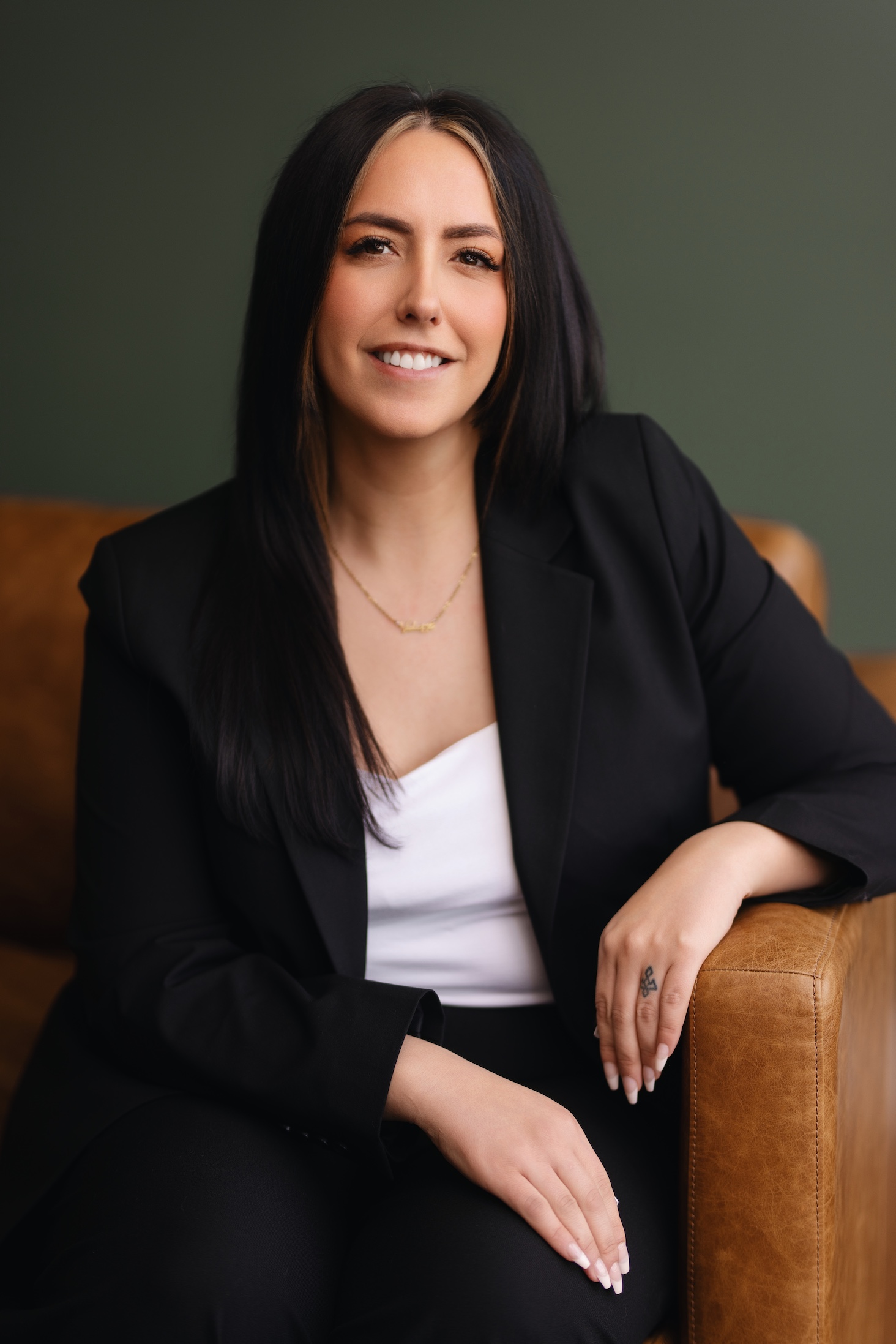 A woman in a black suit smiles while seated on a brown leather chair against a green background.