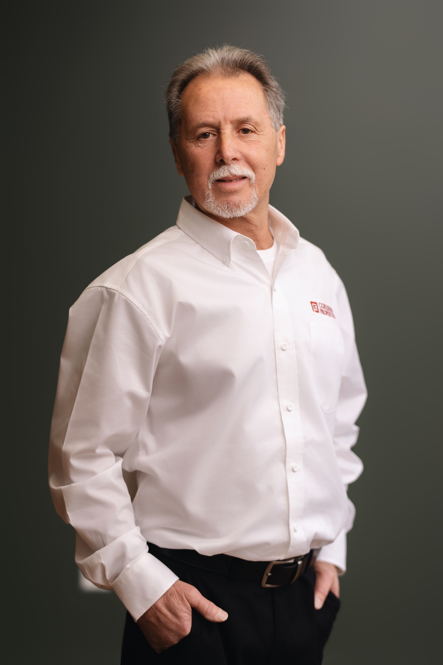 A man with gray hair stands confidently, wearing a white shirt and black pants, hands in pockets.