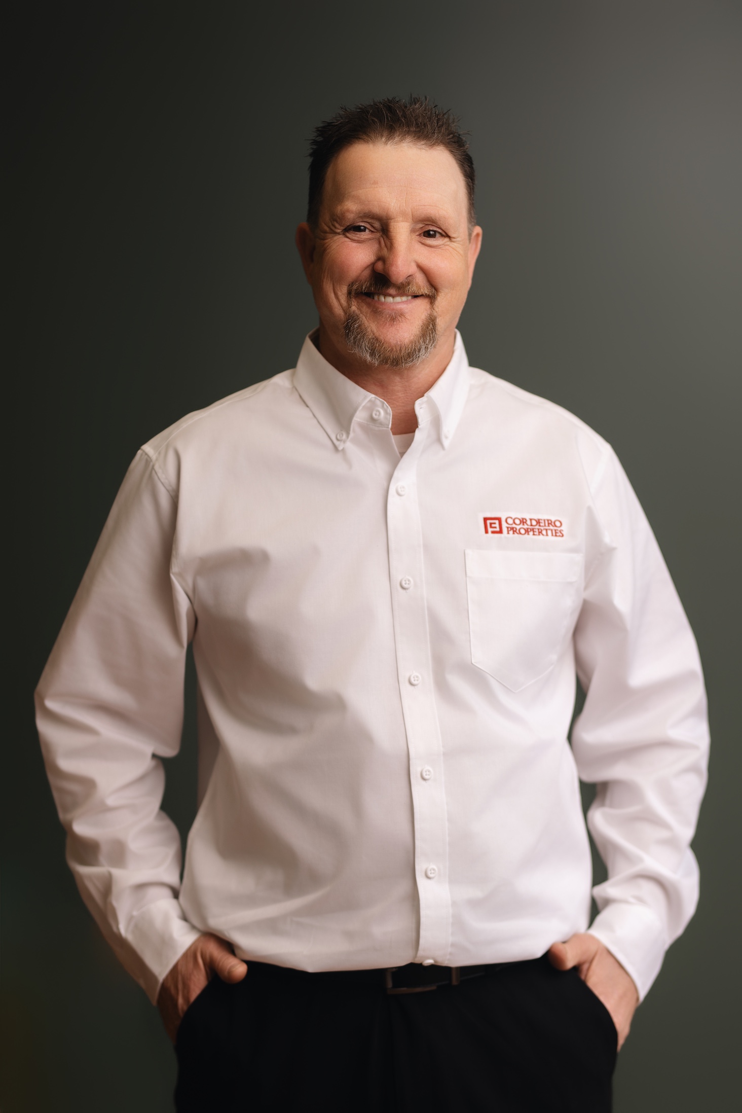 A smiling man in a white shirt with a logo stands with hands in pockets against a dark background.
