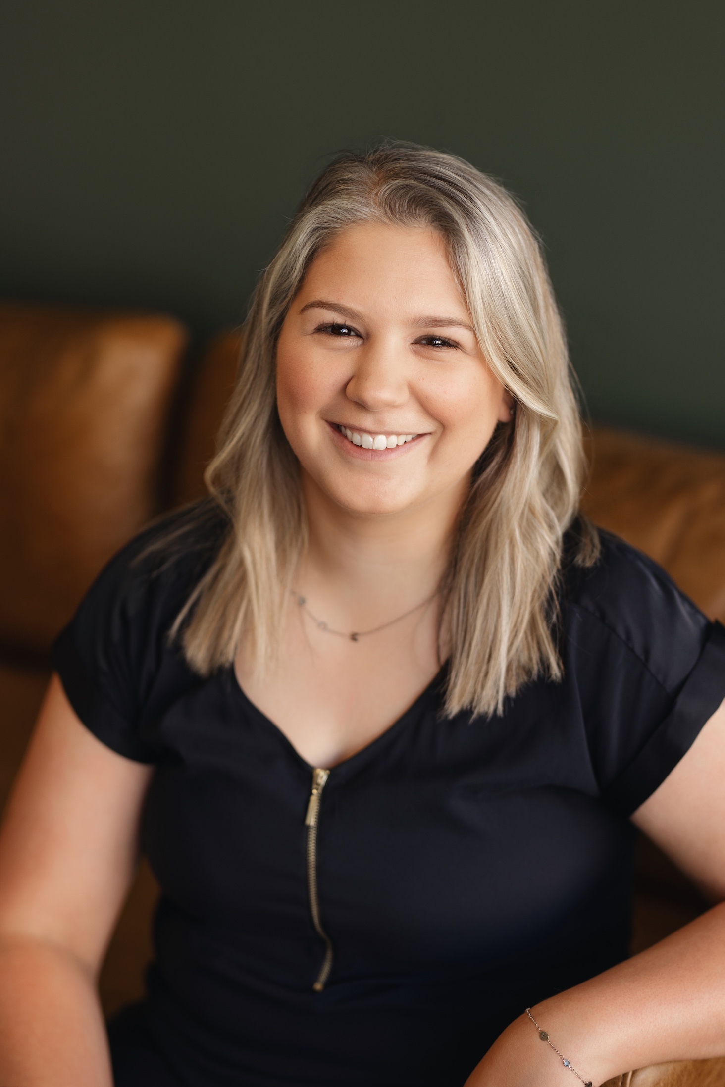 A smiling woman with long, light hair sits on a brown couch, wearing a black top and delicate jewelry.