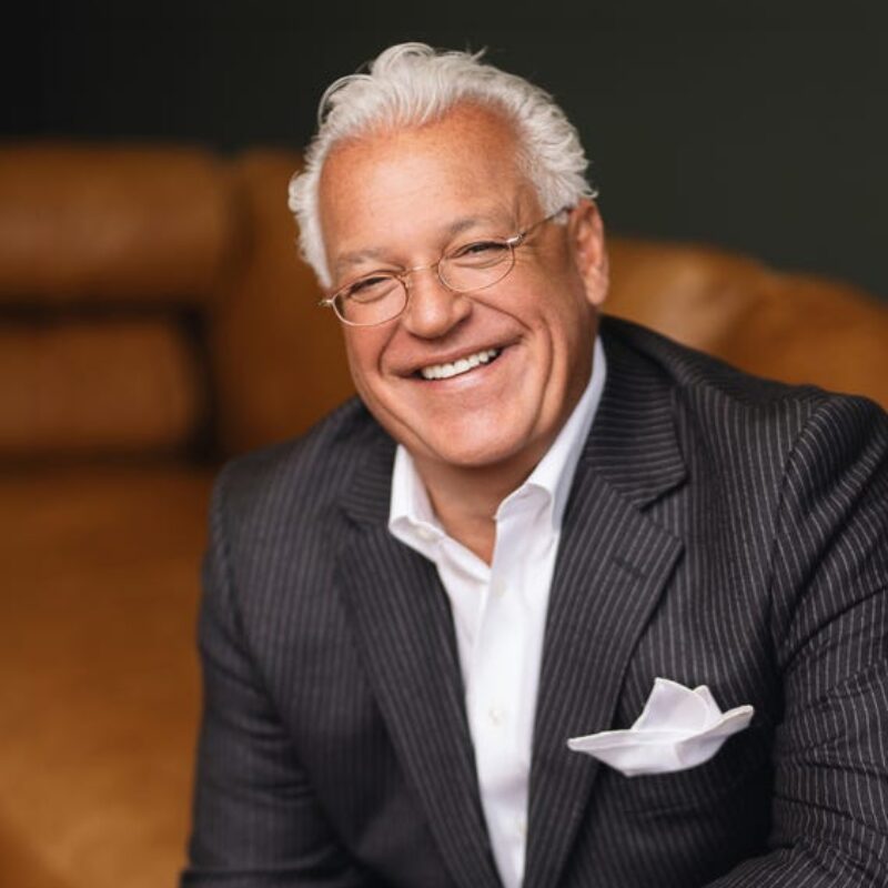 Smiling man in a pinstripe suit sits on a couch, wearing glasses and a white pocket square.