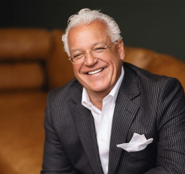 Smiling man in a pinstripe suit sits on a couch, wearing glasses and a white pocket square.